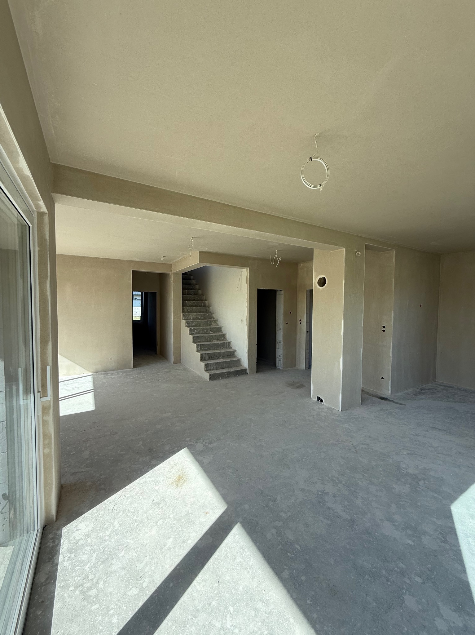 Jasne wnętrze w stanie deweloperskim po nałożeniu tynków, z widocznymi schodami i promieniami słońca wpadającymi przez okno. Na suficie widoczne kable.