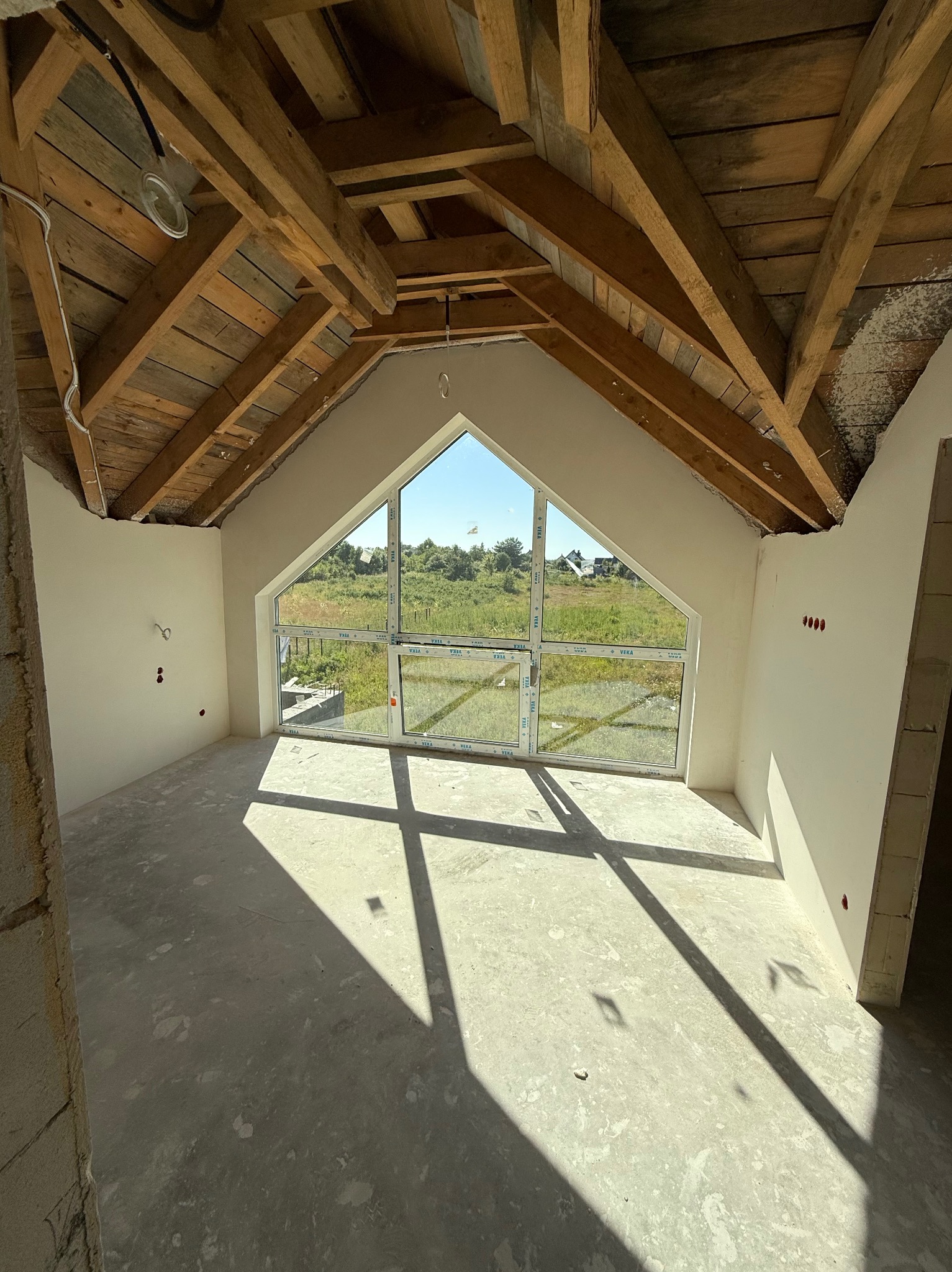 Wnętrze poddasza z drewnianą konstrukcją dachu i oknem szczytowym. Ściany po tynkowaniu, widoczne instalacje elektryczne. Na podłodze cień od okna.