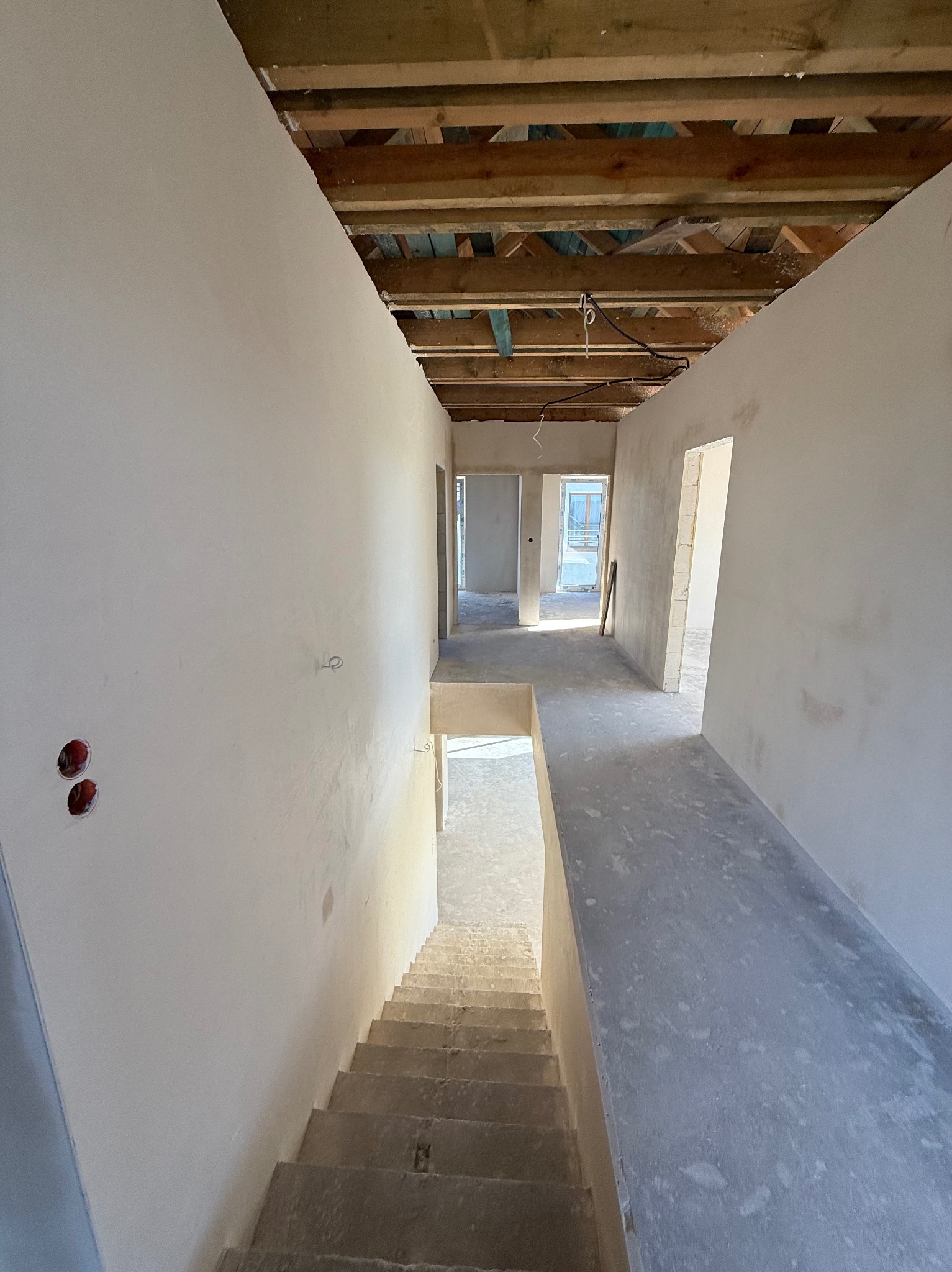 Wnętrze w trakcie prac: świeżo otynkowane ściany w jasnym odcieniu, surowy strop z drewnianych belek i betonowe schody w domu jednorodzinnym.