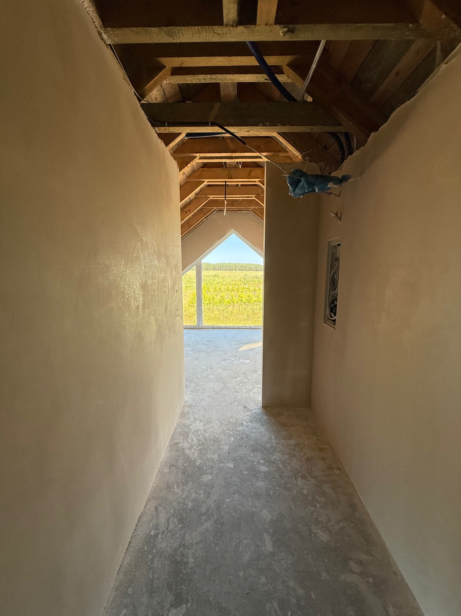 Wnętrze po tynkowaniu maszynowym. Ściany pokryte gładzią, widoczna surowa podłoga i drewniana konstrukcja dachu. W oddali okno z widokiem na pole.