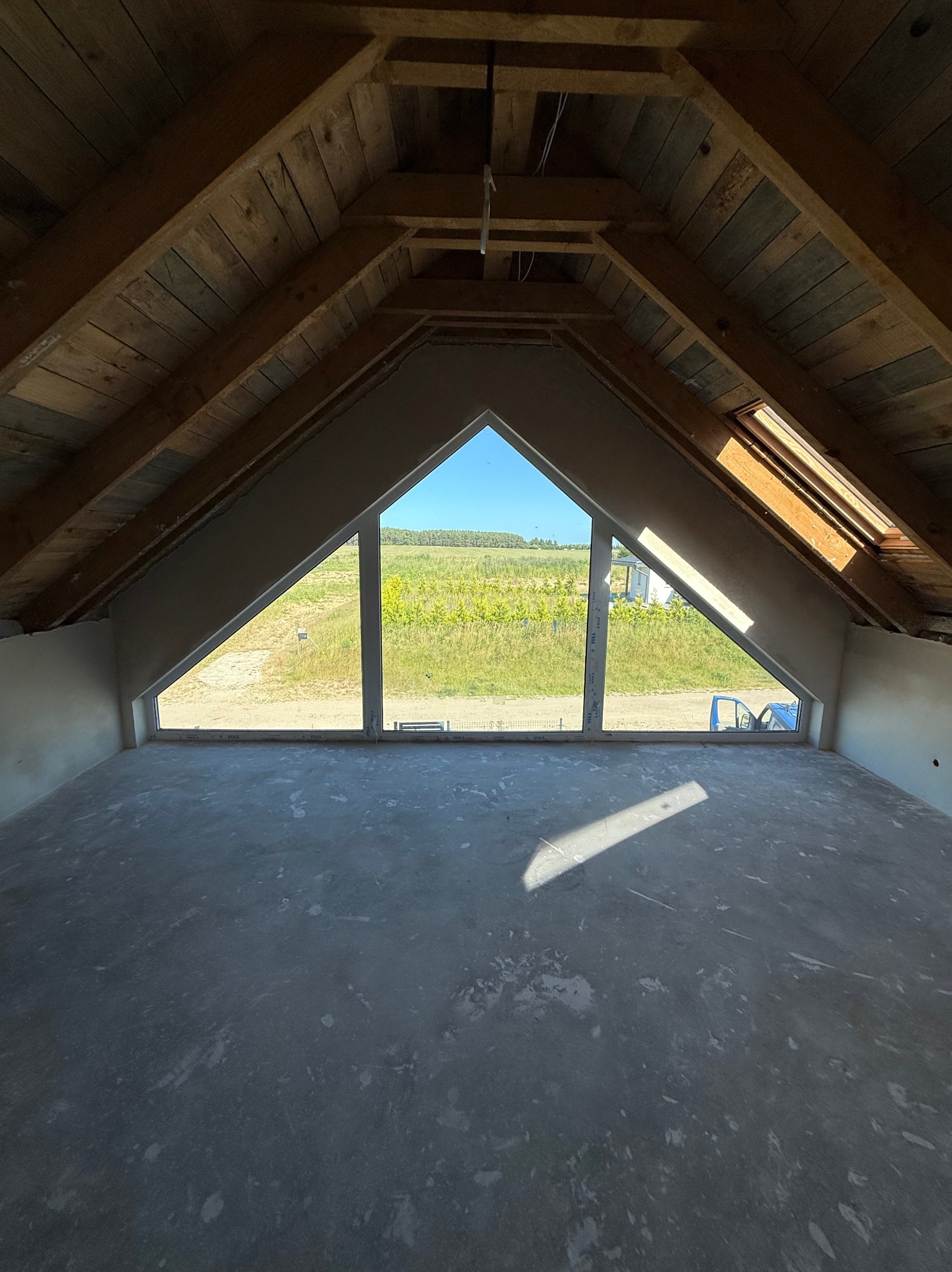 Surowe poddasze z oknem szczytowym i widoczną drewnianą konstrukcją dachu. Ściany częściowo otynkowane, posadzka betonowa, promień słońca na podłodze.
