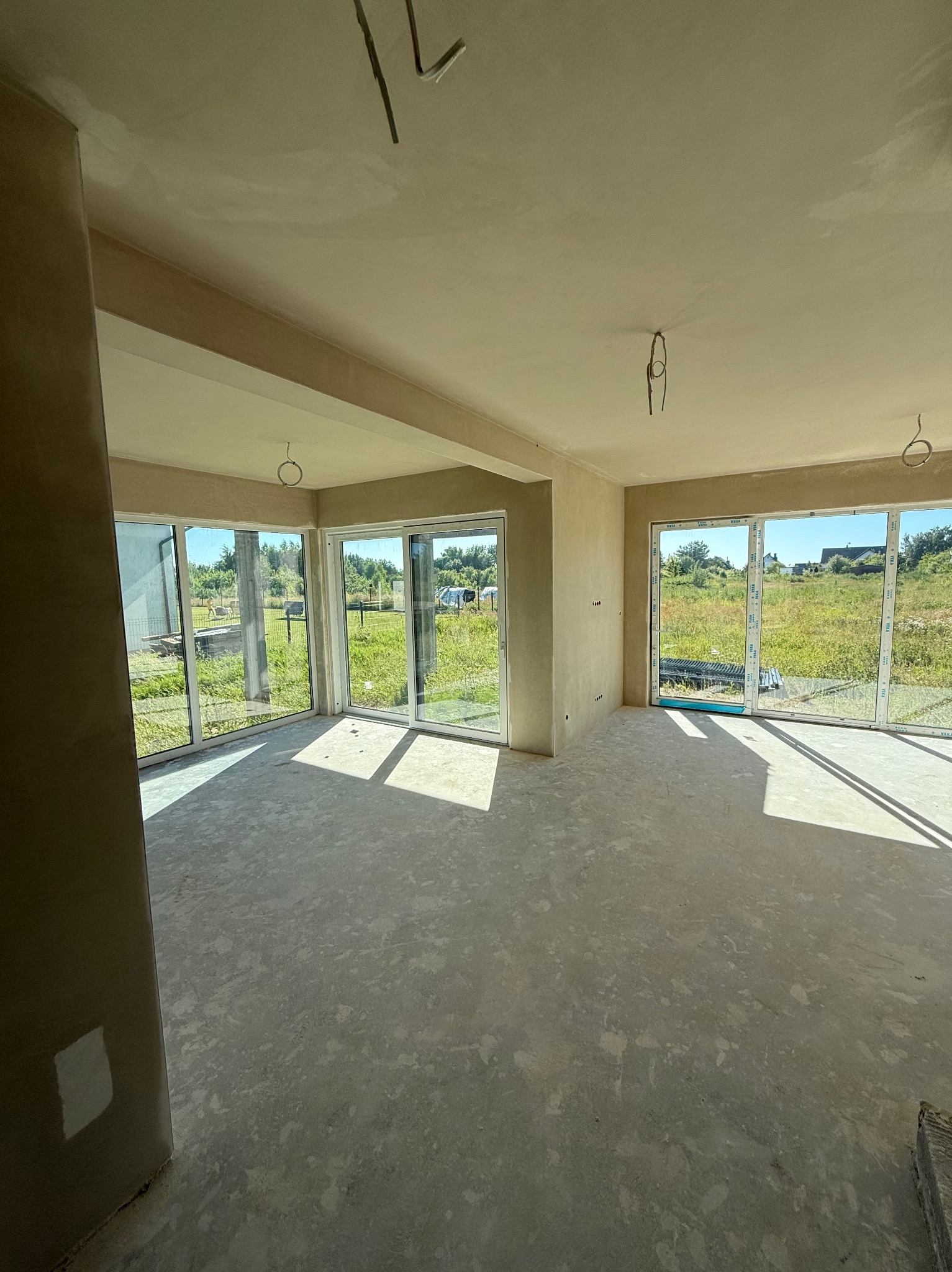 Jasne wnętrze po nałożeniu tynków, z dużymi oknami wychodzącymi na zieleń. Widoczne przewody elektryczne wystające z sufitu i ślady po łatach na ścianie.