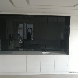 WEGA MEBLE - ALUVITRUM - Nowoczesna kuchnia z białymi szafkami i czarnym panelem szklanym na ścianie, w którym odbija się osoba robiąca zdjęcie i drabina.