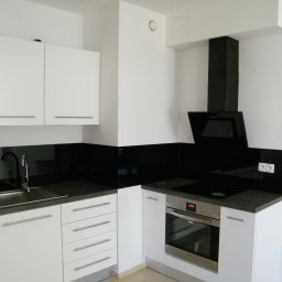 WEGA RAFAŁ ZYCH - Nowoczesna kuchnia w minimalistycznym stylu: białe szafki z poziomymi uchwytami, czarny panel szklany nad blatem, stalowy zlew i bateria, piekarnik oraz czarny okap.