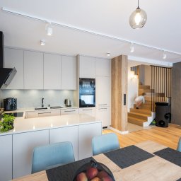 WEGA MEBLE - ALUVITRUM - Nowoczesna kuchnia z jasnymi, lakierowanymi szafkami, wyspą i drewnianymi akcentami, widok na schody z dzieckiem.