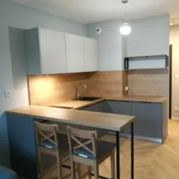 Nowoczesna kuchnia z szarymi szafkami i drewnianym blatem, oświetleniem LED pod szafkami wiszącymi i dwoma drewnianymi hokerami z szarymi poduszkami przy wyspie.