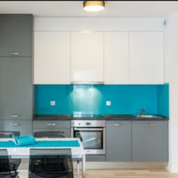 Nowoczesna kuchnia z białymi i szarymi szafkami, turkusowym panelem szklanym nad blatem, oraz stołem z przezroczystymi krzesłami.