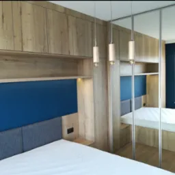 Szafa na wymiar z lustrzanymi drzwiami przesuwnymi, zabudowa nad łóżkiem z drewnianymi szafkami, oświetlenie punktowe i niebieski zagłówek.