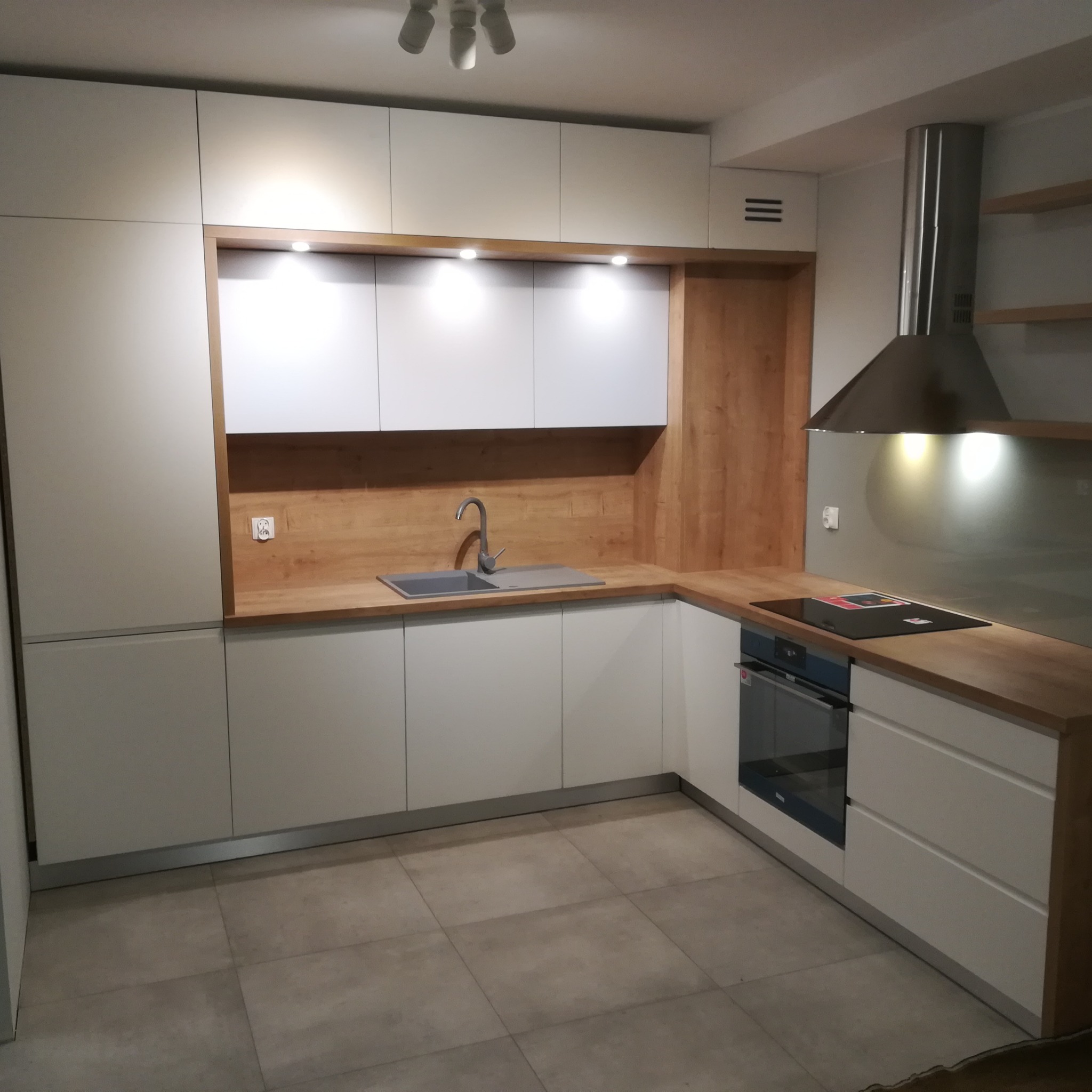 Nowoczesna kuchnia z białymi szafkami i drewnianymi blatami, oświetleniem punktowym pod szafkami wiszącymi i stalowym okapem.