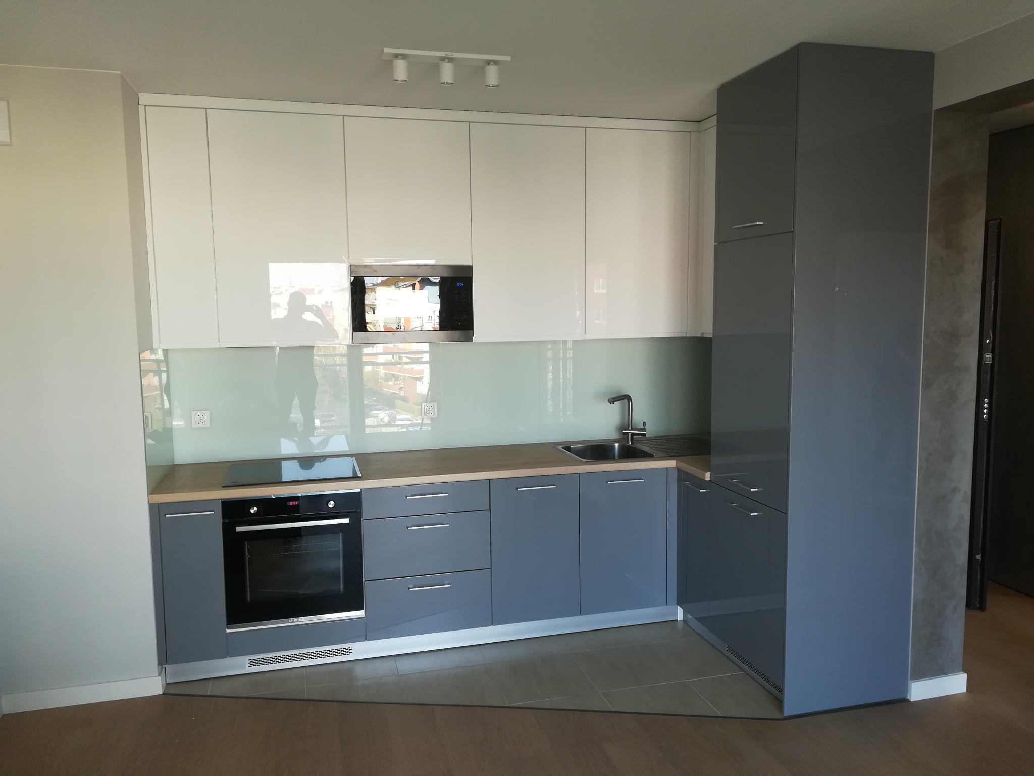 Nowoczesna kuchnia z białymi górnymi szafkami i szarymi dolnymi, z wbudowanym piekarnikiem i mikrofalówką oraz przeszklonym panelem nad blatem.