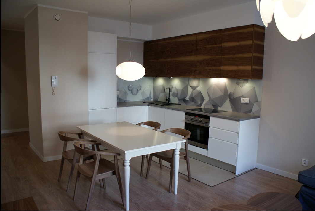 Nowoczesna kuchnia z białymi szafkami, drewnianymi frontami górnymi i geometrycznym wzorem na szklanym panelu nad blatem, oświetlona wiszącą lampą kulą.