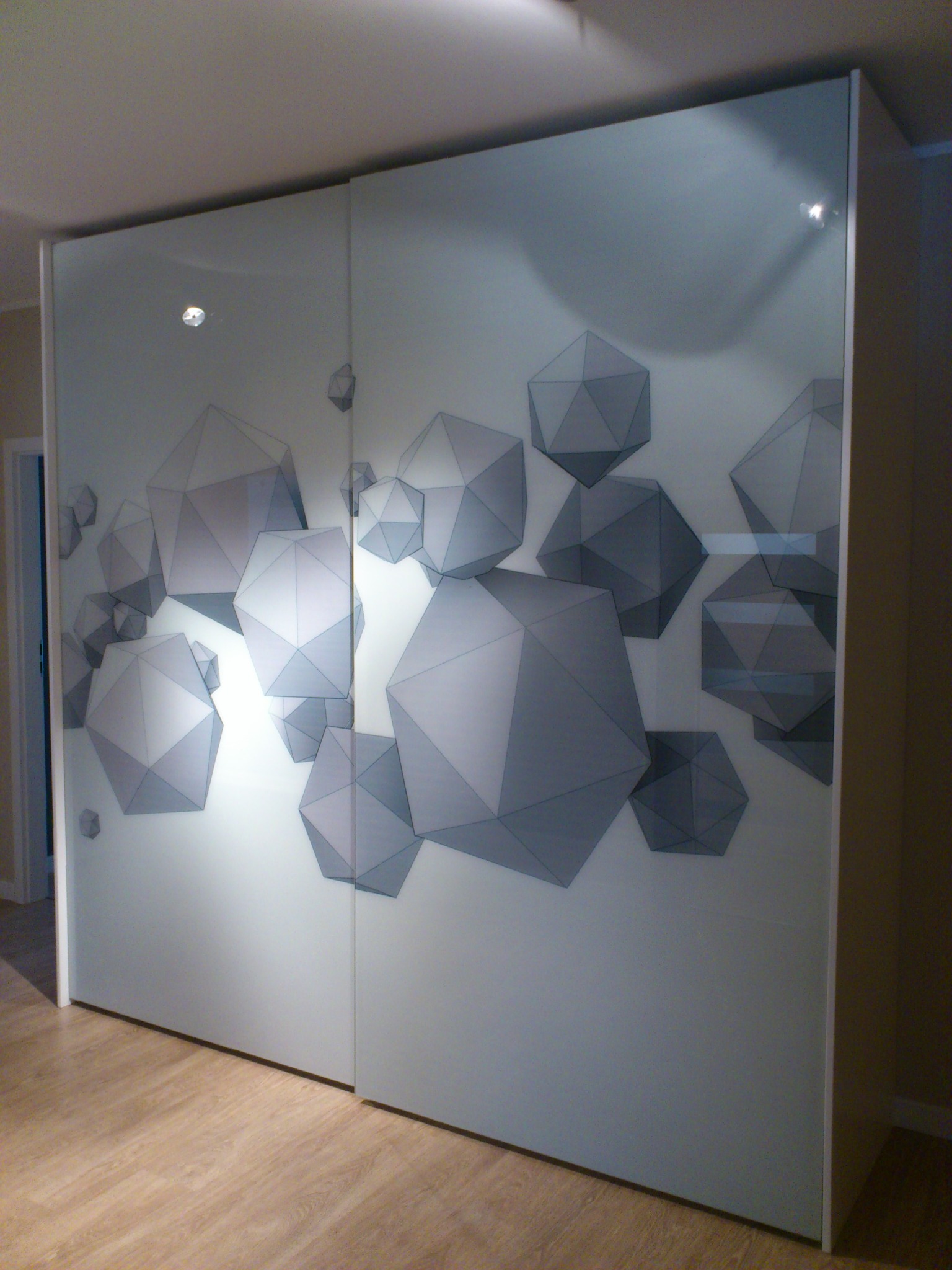 Szafa przesuwna z białymi, matowymi frontami zdobiona motywem szarych brył geometrycznych w przestrzeni mieszkalnej.