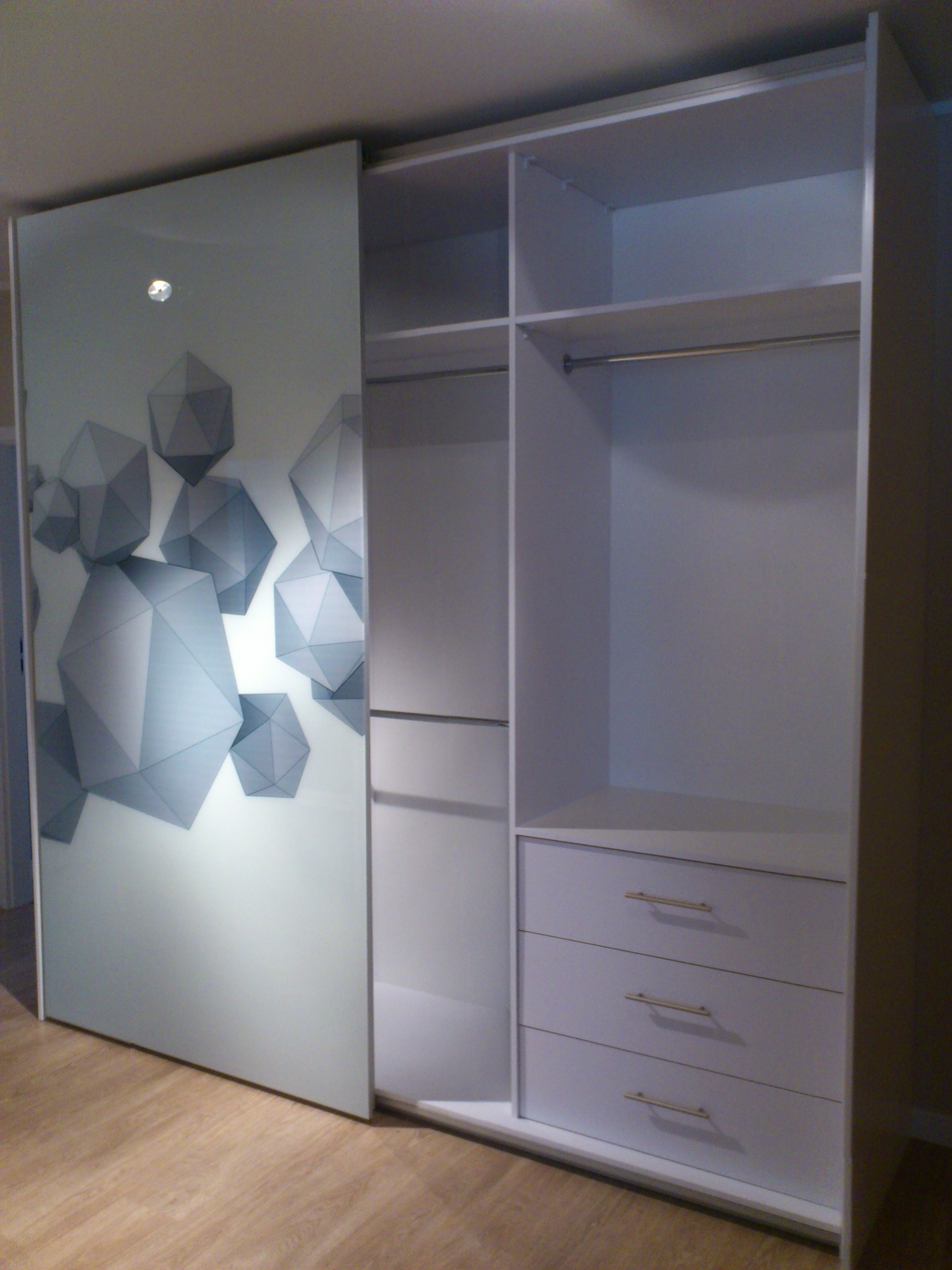 Biała szafa na wymiar z przesuwnymi drzwiami, na których widnieje grafika z szarymi bryłami geometrycznymi, wnętrze szafy z półkami, drążkiem na ubrania i trzema szufladami z podłużnymi, złotymi...