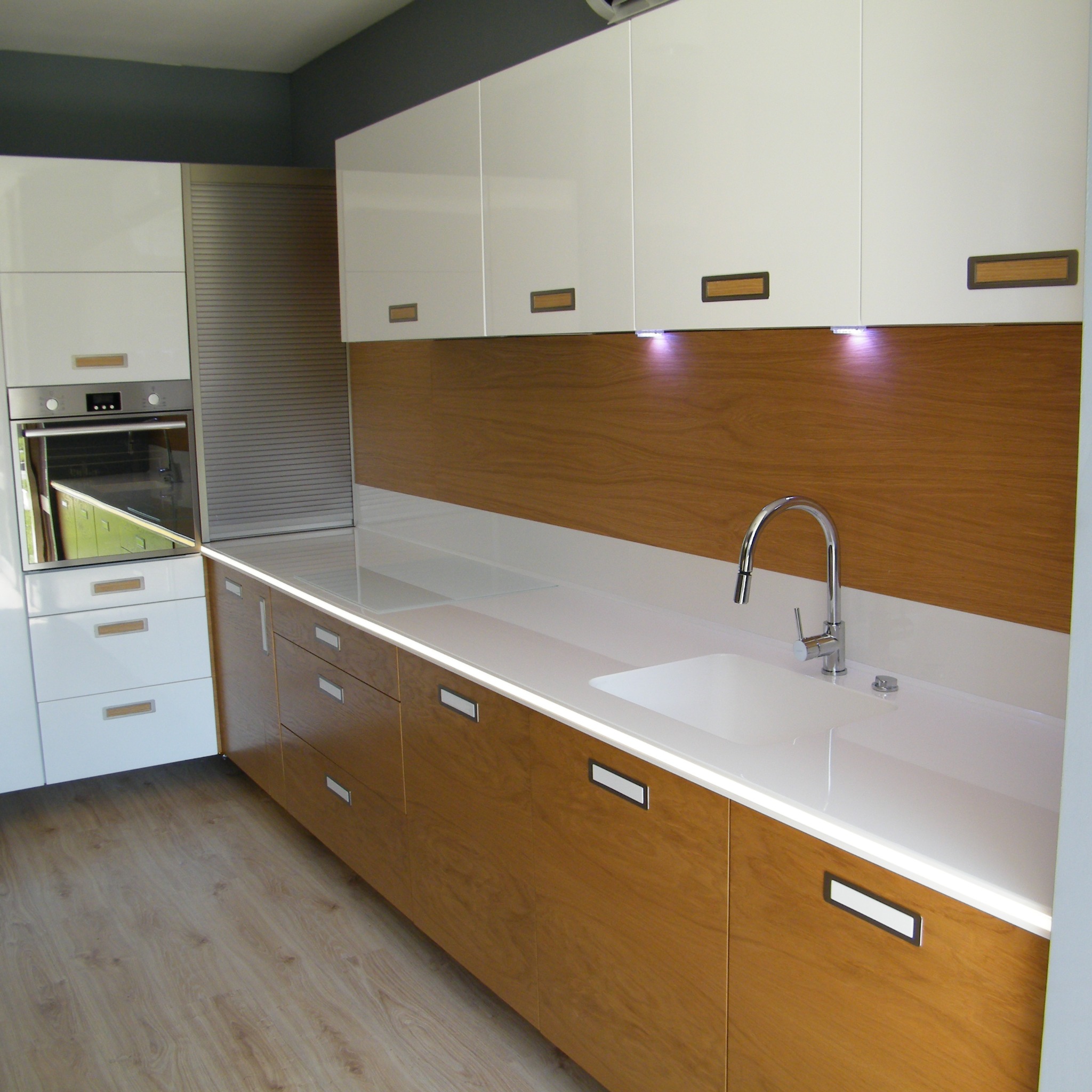 Nowoczesna kuchnia z białymi szafkami górnymi i drewnianymi szafkami dolnymi, oświetlona listwą LED pod blatem, z chromowaną baterią przy białym zlewie.