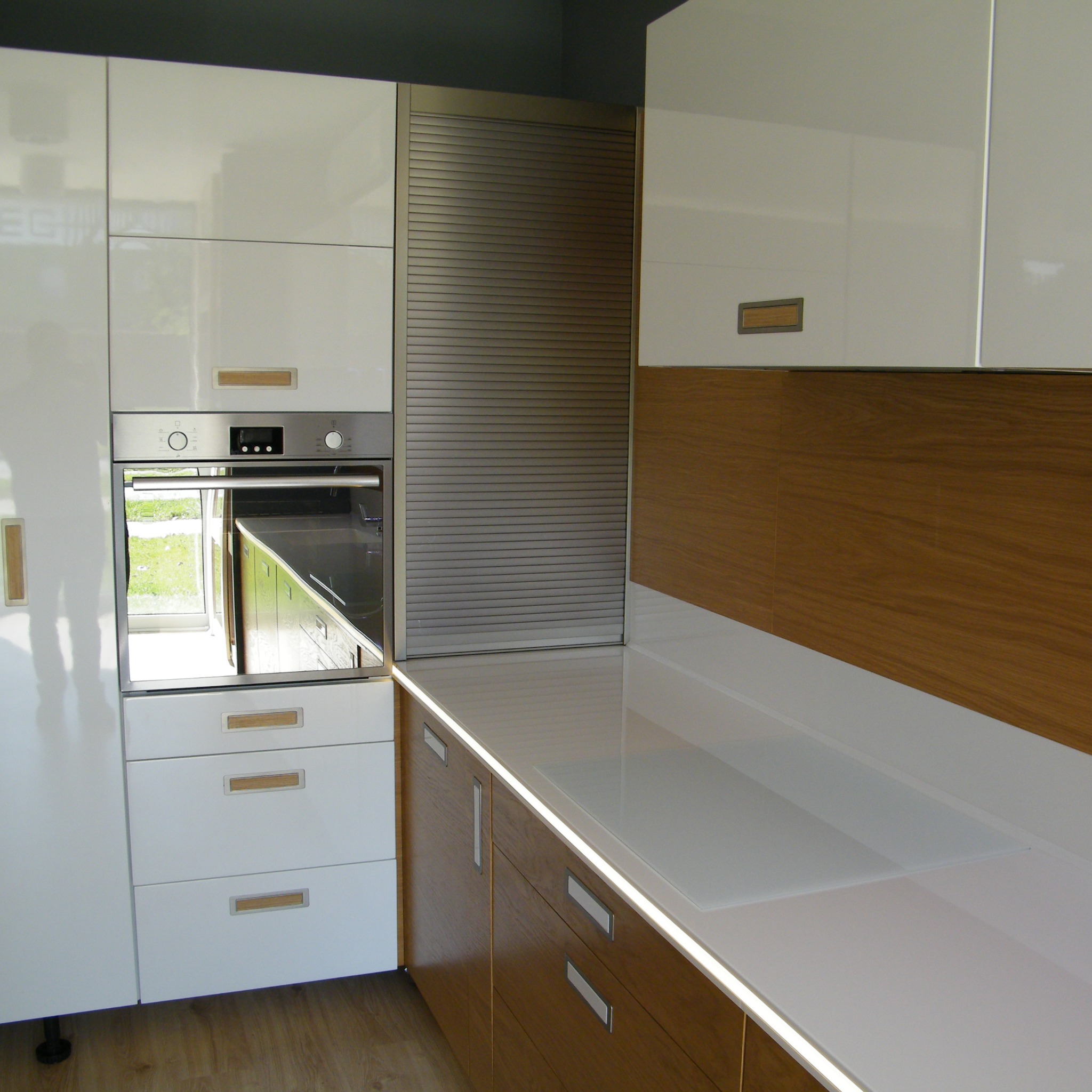 Nowoczesna kuchnia z białymi lakierowanymi szafkami, drewnianymi akcentami i roletą w szafce, oświetlenie LED pod blatem.