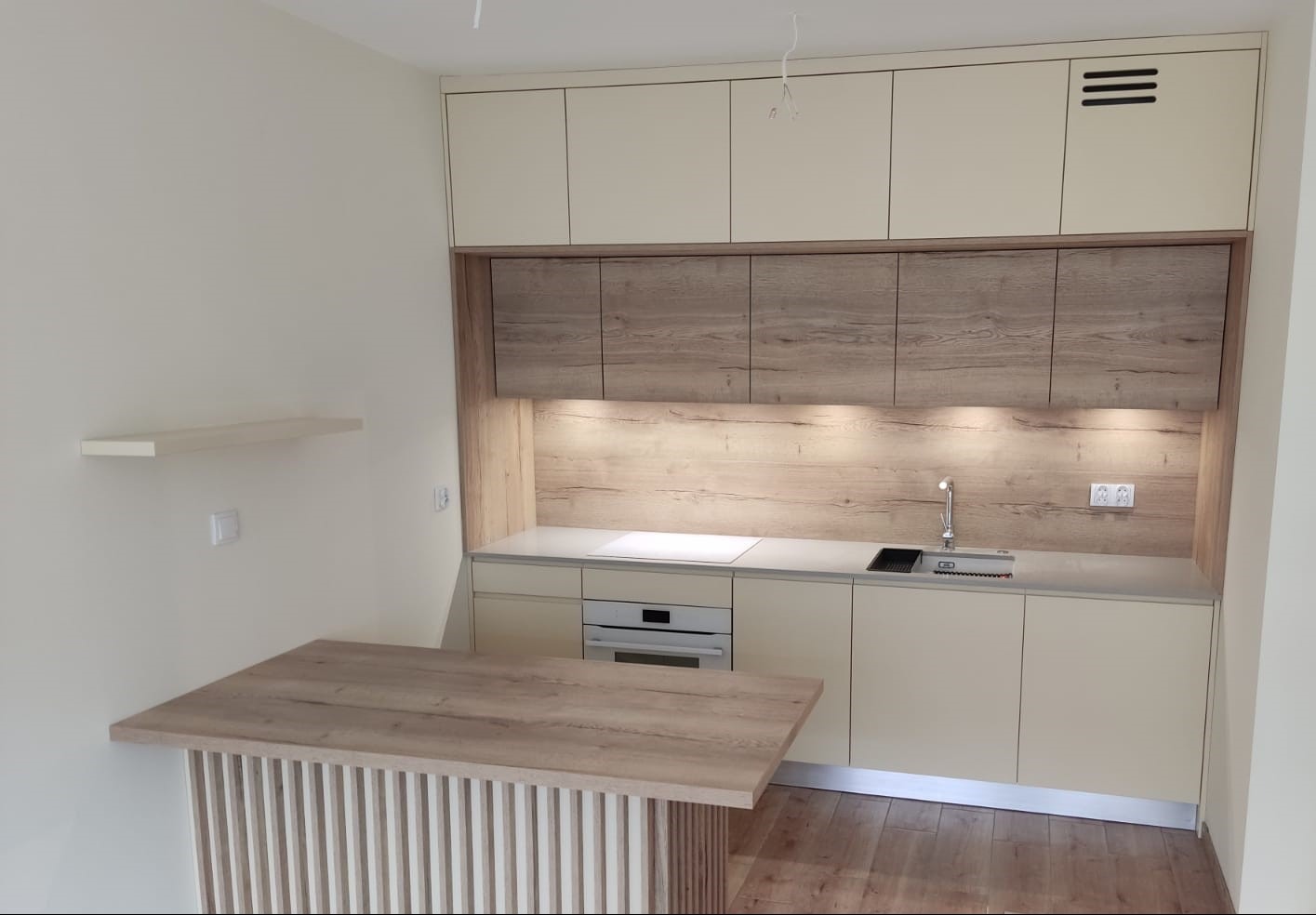 Jasna kuchnia z zabudową meblową na wymiar, blaty w kolorze szarym, drewnopodobne panele ścienne i wyspa z pionowych listewek.