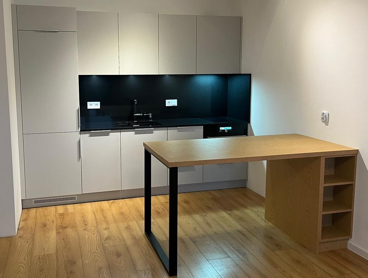 Nowoczesna kuchnia z jasnymi szafkami, czarnym panelem nad blatem i drewnianym stołem z czarnymi nogami, widok na podłogę z paneli.