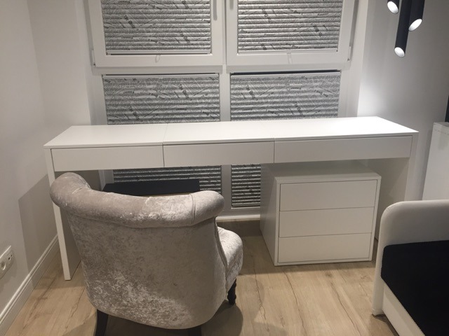 Białe biurko z szufladami i szafką pod oknem z plisowanymi roletami, obok tapicerowane krzesło w kolorze beżowym.