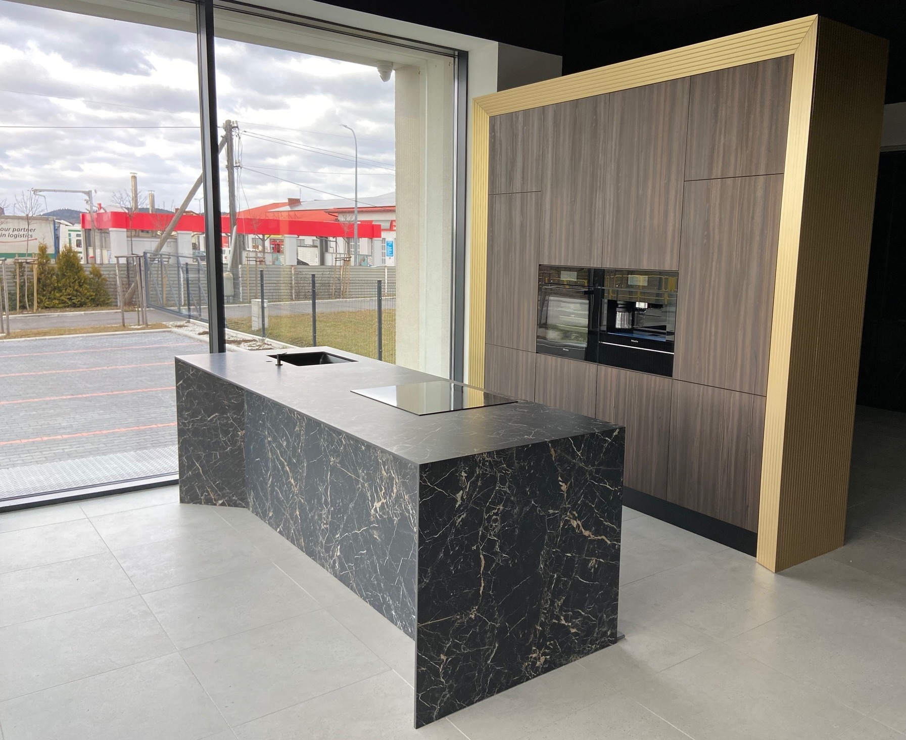 Nowoczesna wyspa kuchenna z czarnego marmuru z wbudowaną płytą indukcyjną i zlewem, na tle zabudowy kuchennej z ciemnego drewna z elementami w kolorze złota i wbudowanymi urządzeniami AGD.