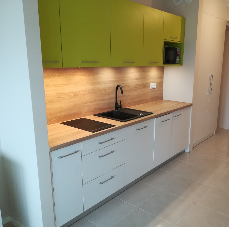 Nowoczesna kuchnia z białymi szafkami dolnymi, zielonymi szafkami górnymi i drewnianym blatem, wyposażona w czarny zlew i płytę indukcyjną.