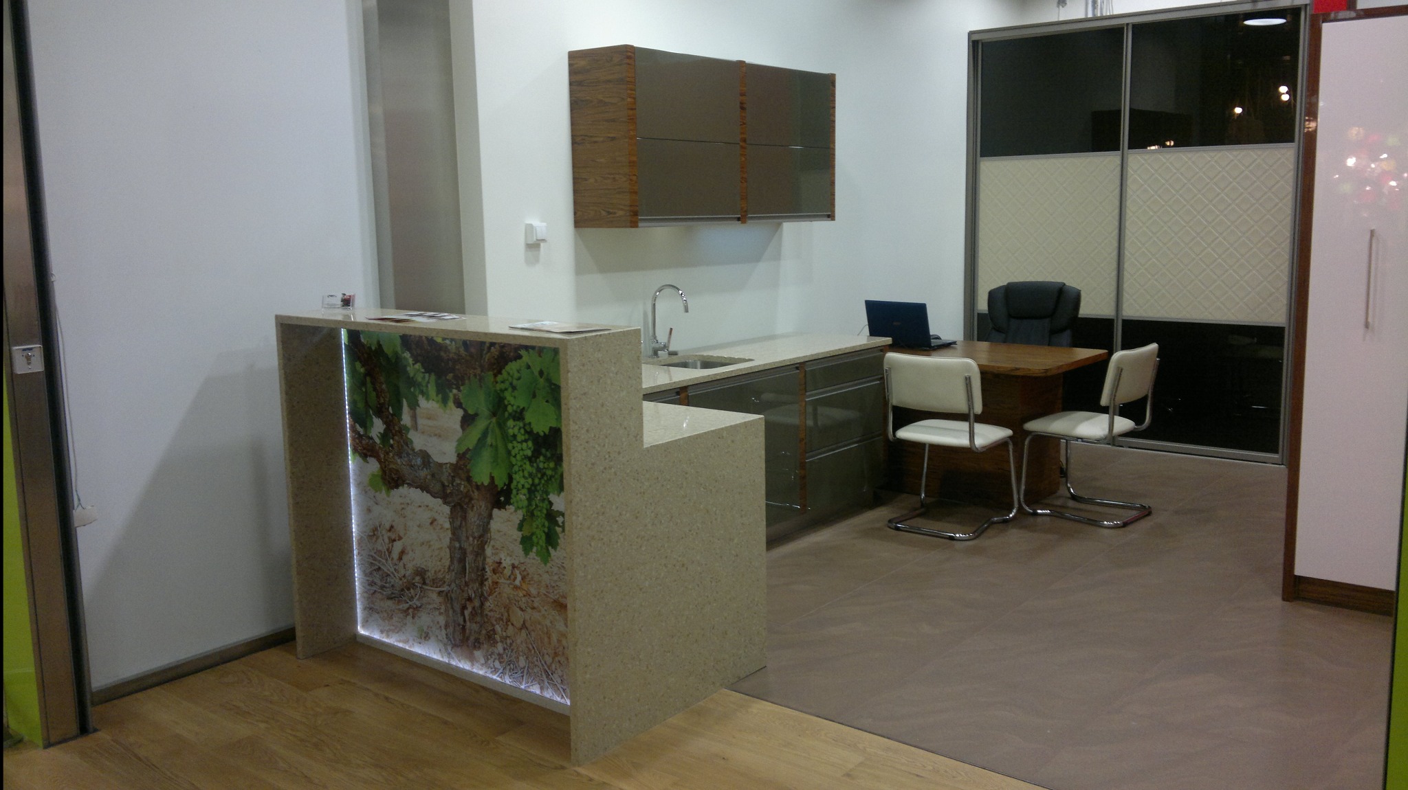 Wnętrze biura z recepcją z kamiennego konglomeratu z podświetleniem LED i grafiką winorośli, szafki wiszące w kolorze taupe, przesuwne drzwi szklane z wzorem, drewniany stół i krzesła.