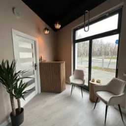 Nowoczesne wnętrze biura z recepcją wykończoną drewnianymi lamelami, dwoma krzesłami obitymi beżowym welurem, stolikiem pomocniczym i dużym oknem z widokiem na ulicę. Na podłodze jasne panele...