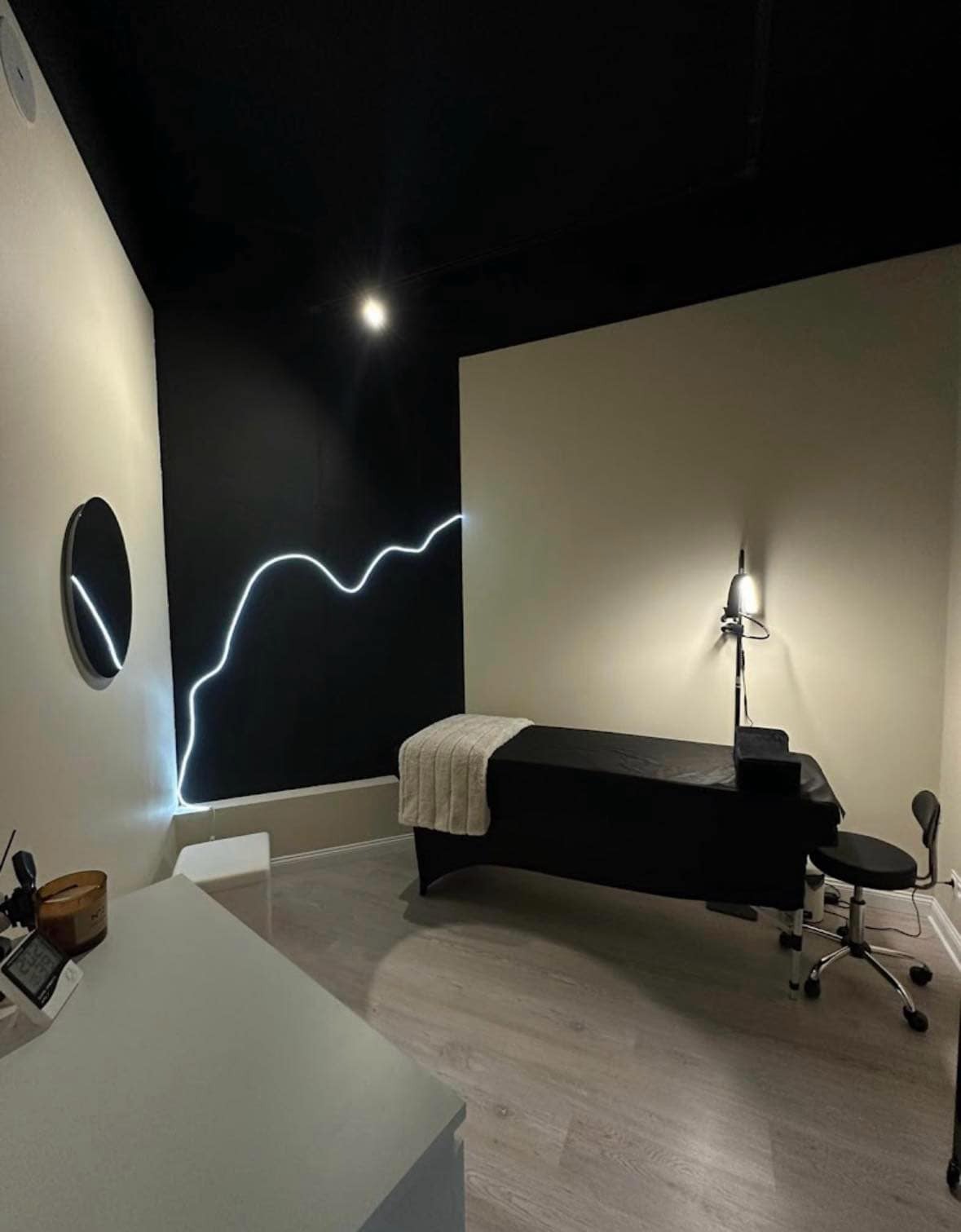 Minimalistyczne wnętrze gabinetu w Tłuszczu z czarnym sufitem i ścianą, oświetlone linią ledową w kształcie fali, z ciemnym łóżkiem do masażu i krzesłem na kółkach.