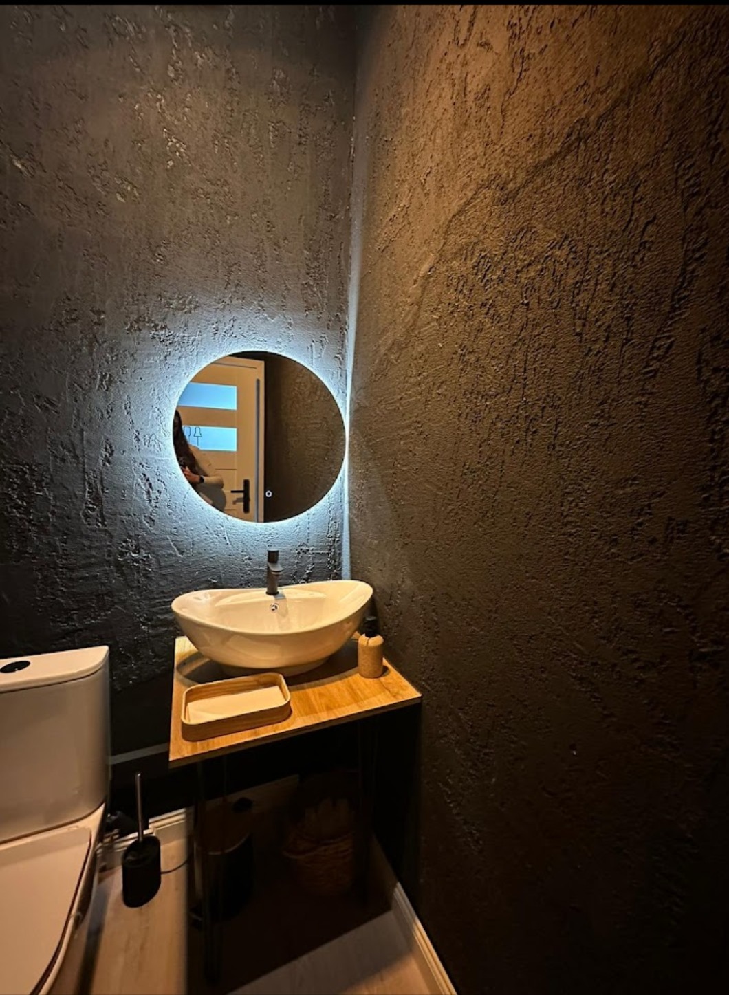Nowoczesna łazienka z umywalką nablatową, okrągłym lustrem z podświetleniem LED i ścianami pokrytymi ciemną, strukturalną farbą.
