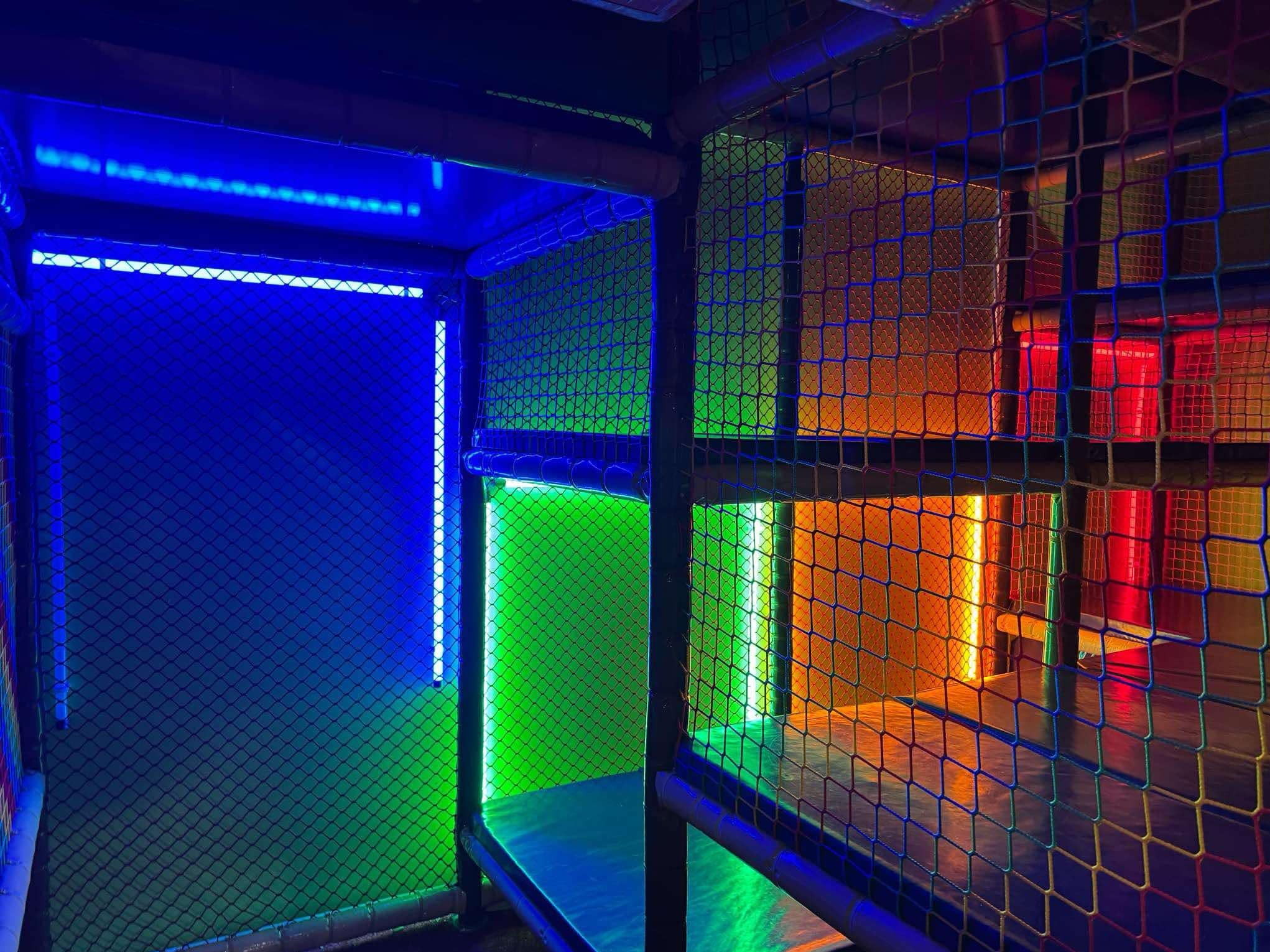 Wielopoziomowa konstrukcja zabawowa z siatki, oświetlona różnokolorowymi diodami LED: niebieskim, zielonym, pomarańczowym i czerwonym.