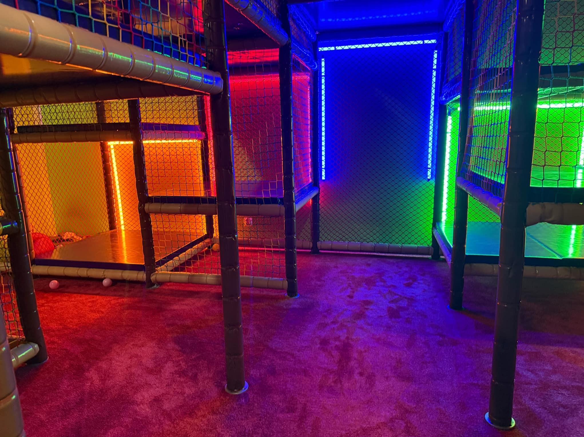 Wnętrze sali zabaw dla dzieci z wielopoziomową konstrukcją z siatkami, oświetloną kolorowymi neonami w odcieniach czerwieni, niebieskiego, zieleni i pomarańczy, z fioletową wykładziną na podłodze...