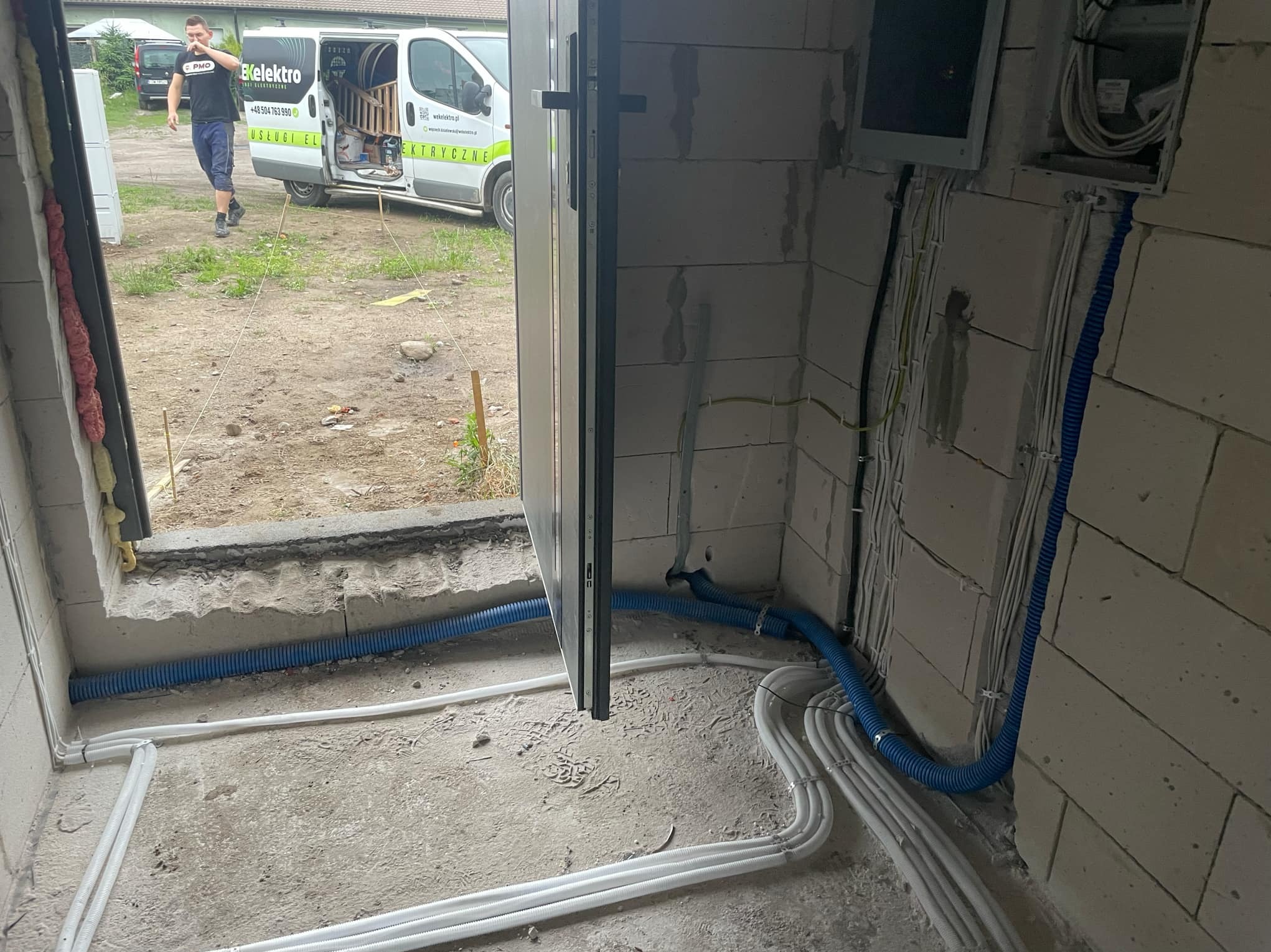 Wnętrze w stanie surowym z widocznymi przewodami elektrycznymi w białych i niebieskich peszlach, układanymi wzdłuż ściany z betonu komórkowego. Na zewnątrz widoczny samochód dostawczy z logo firmy...