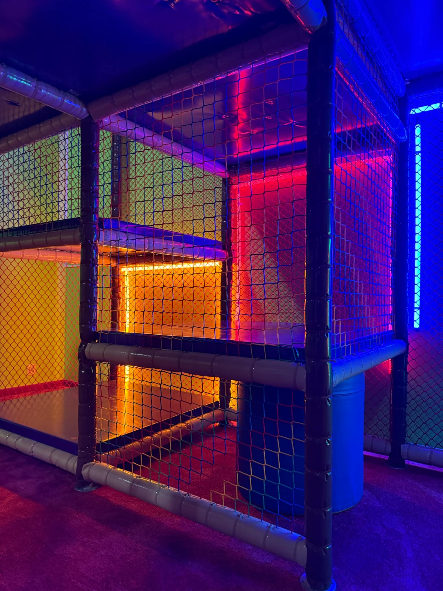 Wielopoziomowa konstrukcja zabawowa z siatki, oświetlona neonowymi światłami w kolorach niebieskim, czerwonym i pomarańczowym, z elementami konstrukcyjnymi z rur PCV.