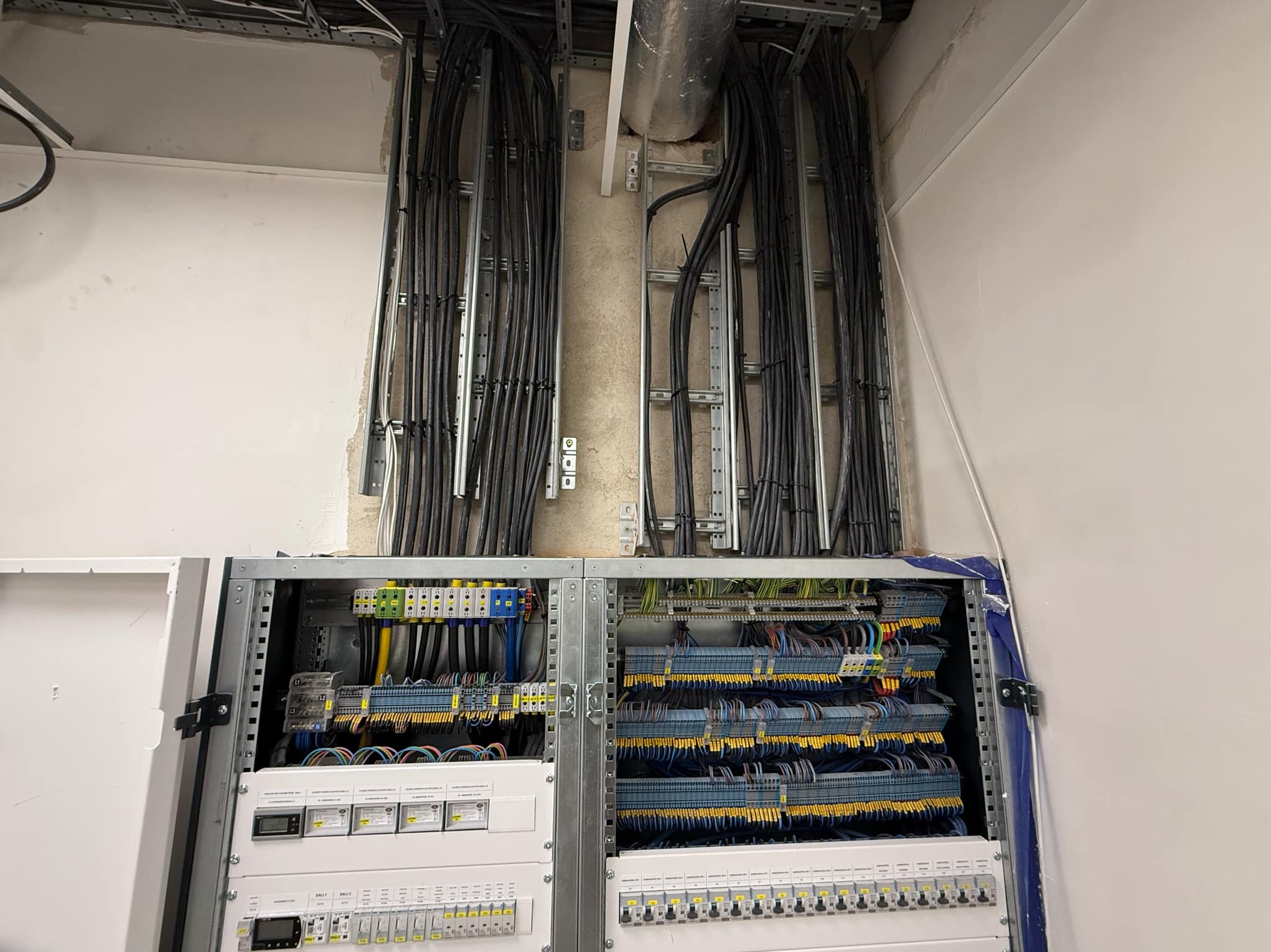 Rozdzielnica elektryczna z licznymi przewodami w korytach kablowych, widoczne zabezpieczenia, moduły i listwy zaciskowe w budynku w stanie deweloperskim.