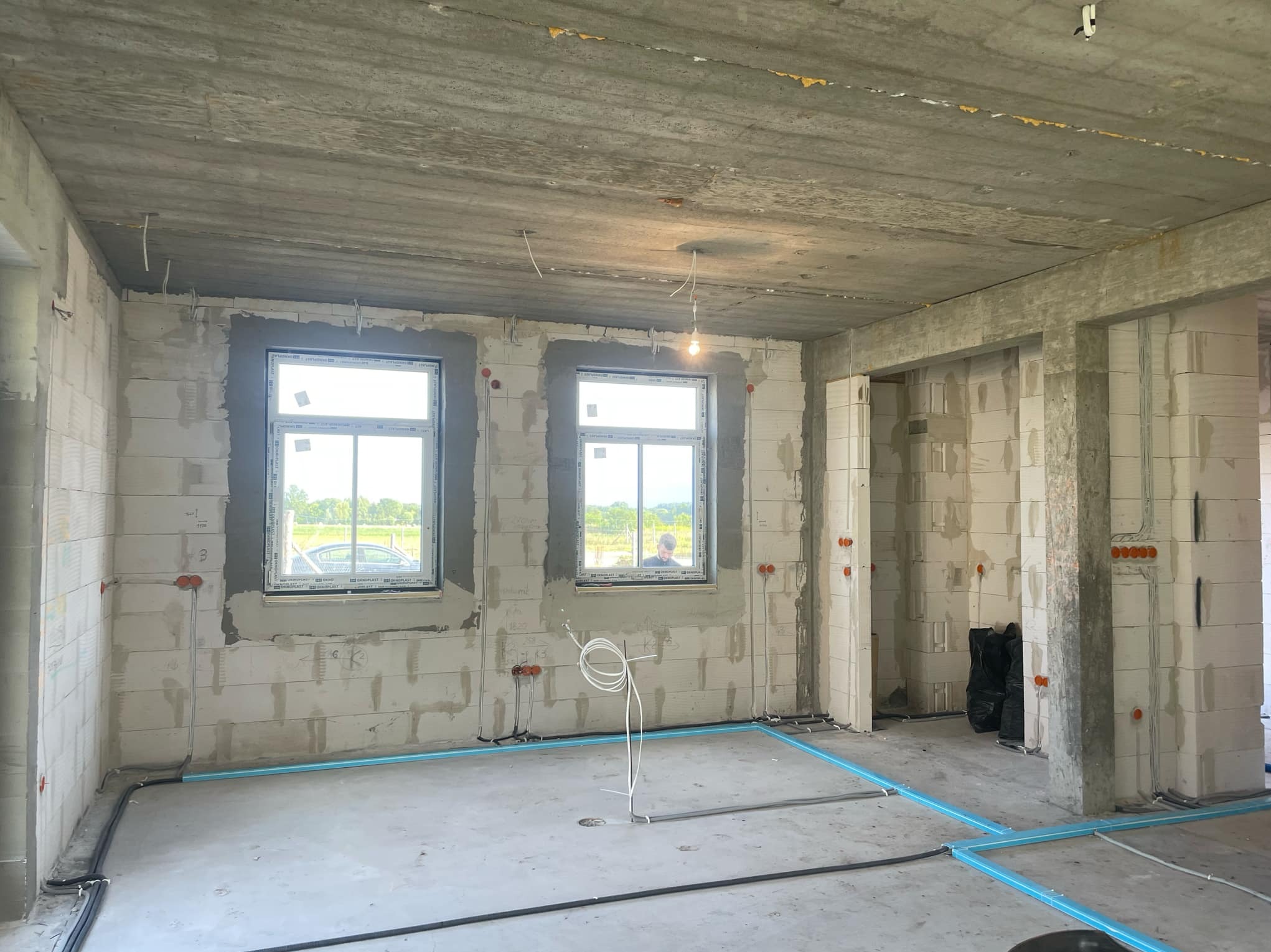 Surowy stan wnętrza z widocznymi przewodami elektrycznymi w ścianach i podłodze, puszki instalacyjne wmurowane w bloczki, tymczasowe oświetlenie żarówką, okna z widokiem na zieleń na zewnątrz.