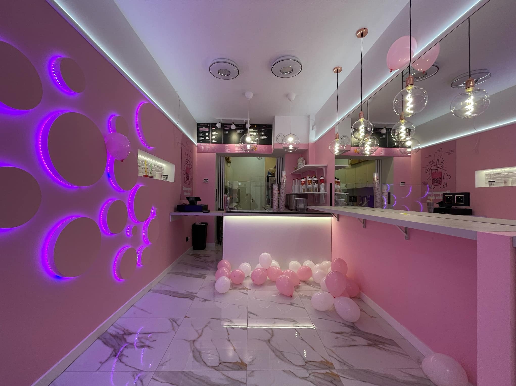 Wnętrze lokalu w odcieniach różu z dekoracyjnym oświetleniem LED w kształcie okręgów na ścianie, wiszącymi lampami z żarówkami Edisona i balonami na podłodze z marmurową fakturą.