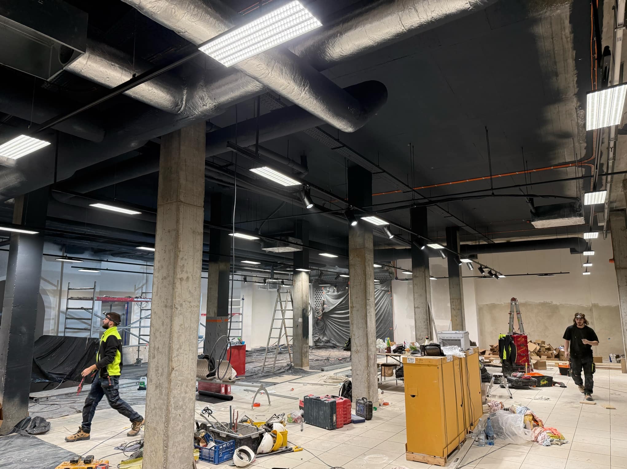Wnętrze w trakcie remontu z widocznymi instalacjami pod sufitem, pracownik w odblaskowej kamizelce i narzędzia porozrzucane na podłodze.