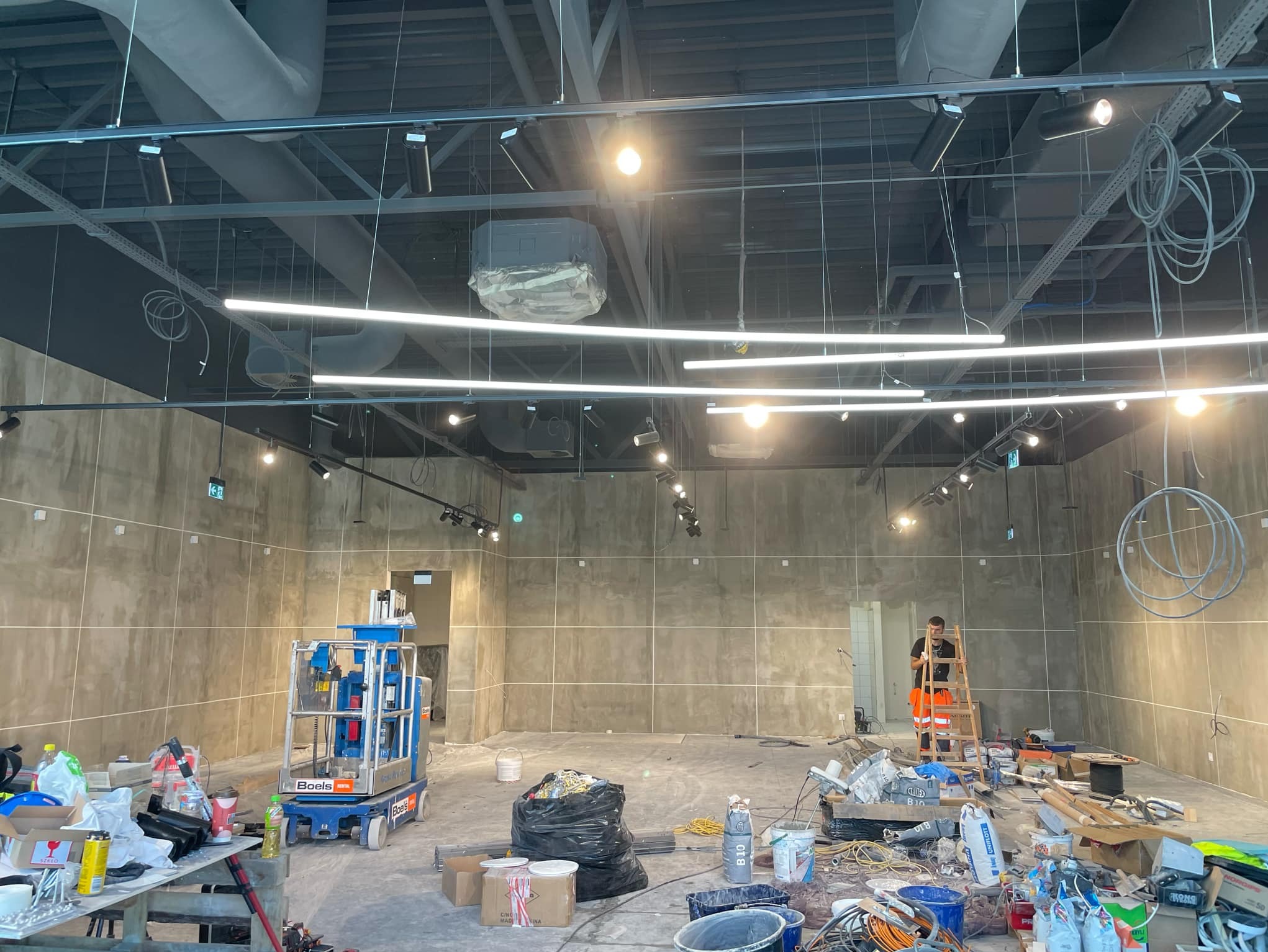 Wnętrze komercyjne w trakcie montażu oświetlenia: widoczne lampy liniowe LED, reflektory szynowe, podnośnik koszowy Boels, drabina z pracownikiem w odblaskowej kamizelce oraz materiały budowlane.