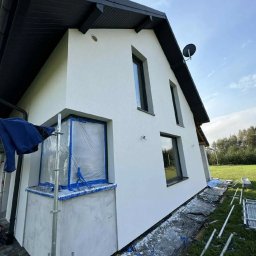 Łukasz Górka Usługi remontowo budowlane - Biała elewacja domu jednorodzinnego po tynkowaniu, z zabezpieczonym oknem folią i rozłożonymi rusztowaniami. Widok z dołu, podkreślający geometrię budynku.