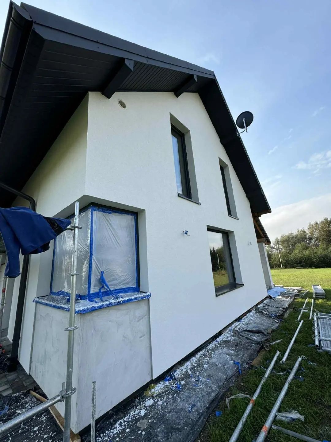 Biała elewacja domu jednorodzinnego po tynkowaniu, z zabezpieczonym oknem folią i rozłożonymi rusztowaniami. Widok z dołu, podkreślający geometrię budynku.