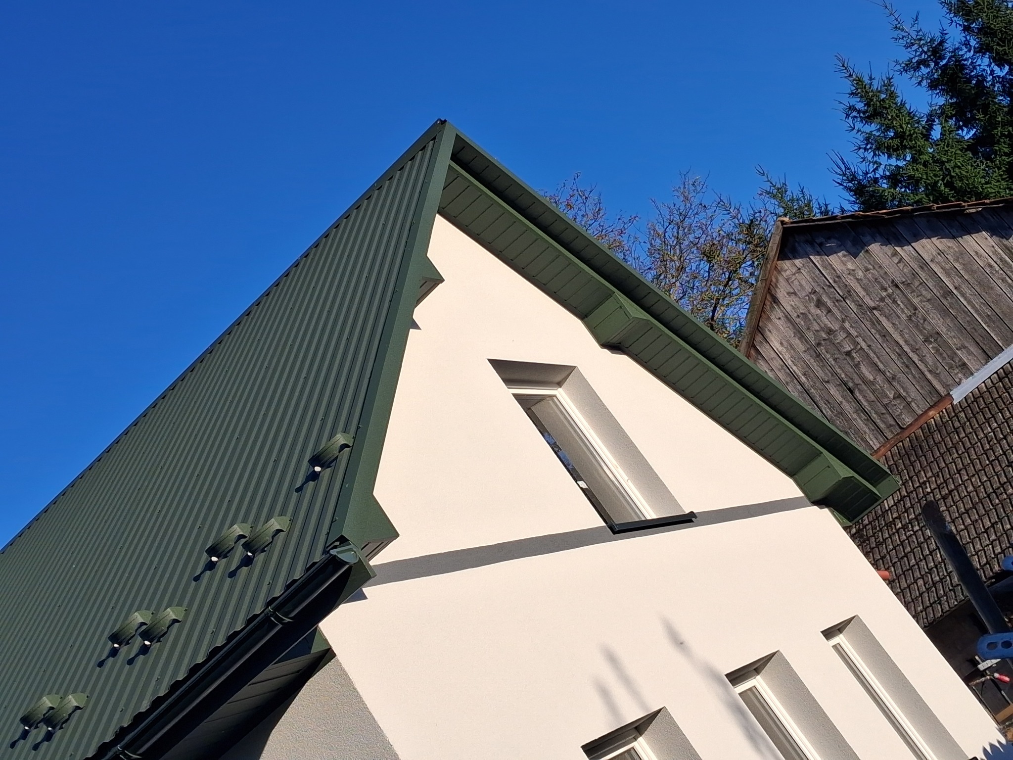 Biała elewacja domu z zielonym dachem z blachy trapezowej, widok z dołu pod kątem, błękitne niebo w tle, fragment drewnianego budynku obok.