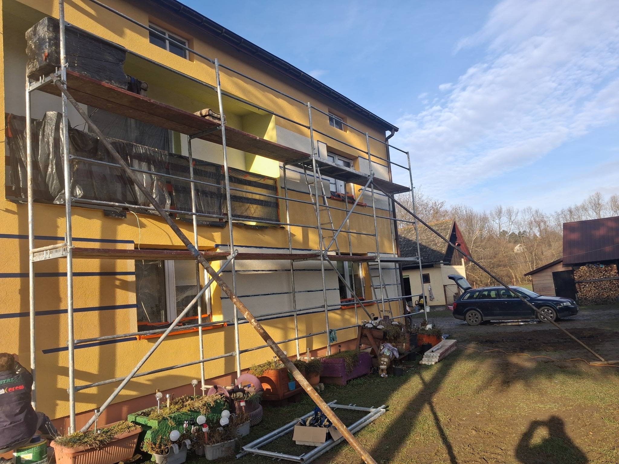Żółty dom w trakcie prac elewacyjnych, z rusztowaniem i widocznymi fragmentami ocieplenia. Na pierwszym planie donice z roślinami i elementy rusztowania.