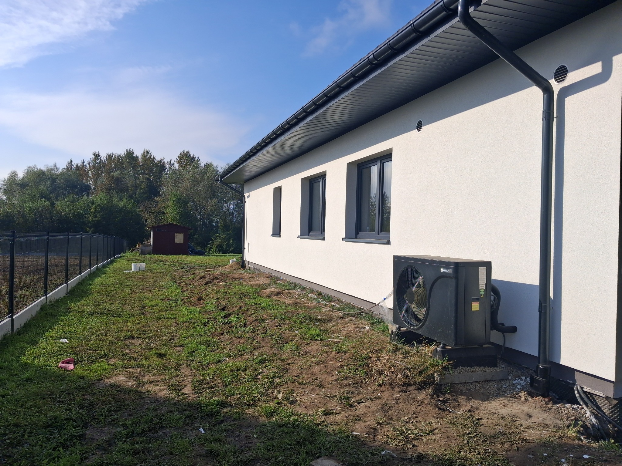 Biała elewacja domu z czarnymi oknami i klimatyzatorem na zewnątrz, widok z boku na trawnik z ogrodzeniem i drzewami w tle, słoneczny dzień.