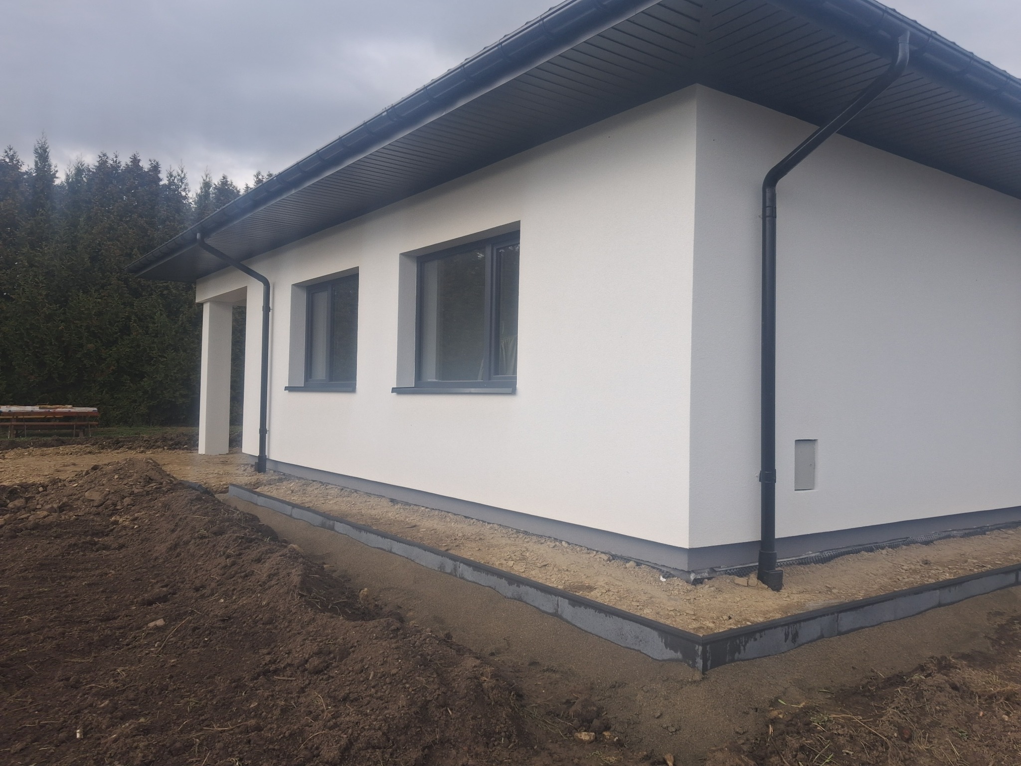 Biały, parterowy dom z szarą stolarką okienną i dachem. Widoczne obrzeża wokół budynku i przygotowany teren pod zagospodarowanie.