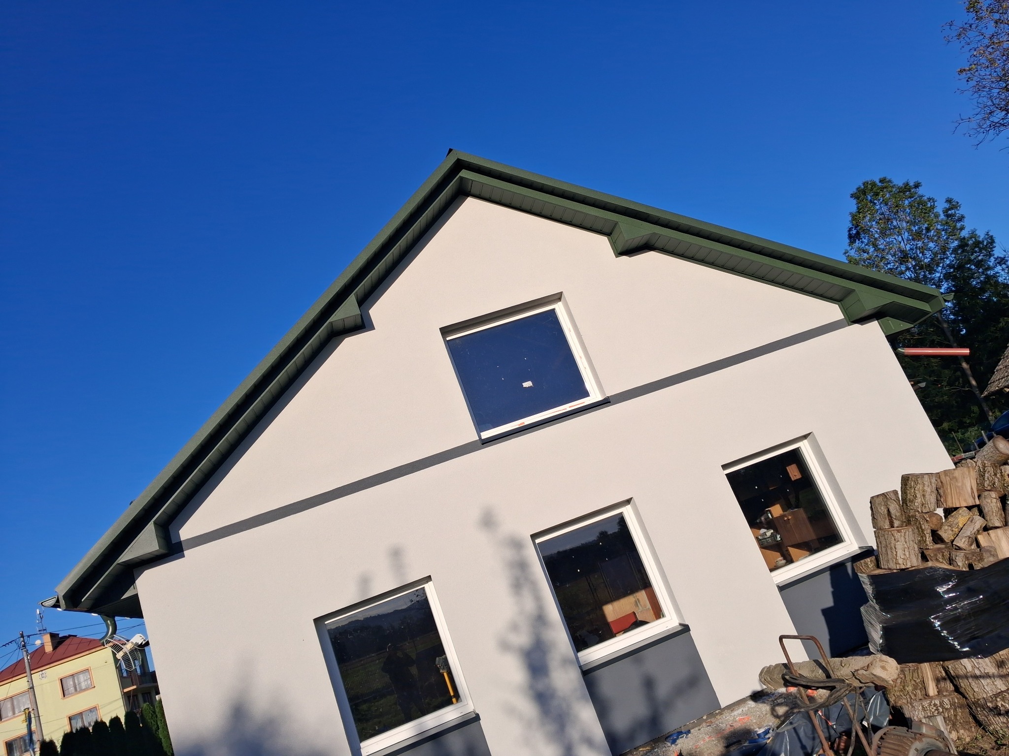 Nowoczesny dom jednorodzinny w Kniażycach z jasną elewacją i zielonym dachem, uchwycony pod kątem w słoneczny dzień. Widoczne okna z odbiciami.