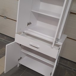 Taboo Piotr Tabor - Biała szafka z otwartymi drzwiczkami i szufladą, widok z boku na tle garażowej bramy. Prosta forma, minimalistyczny design, metalowe uchwyty.