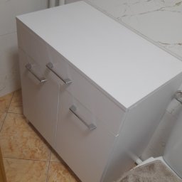 Taboo Piotr Tabor - Biała szafka łazienkowa z dwoma drzwiczkami i srebrnymi uchwytami, stojąca na brązowych płytkach, widok z góry, elementy montażowe na sedesie.