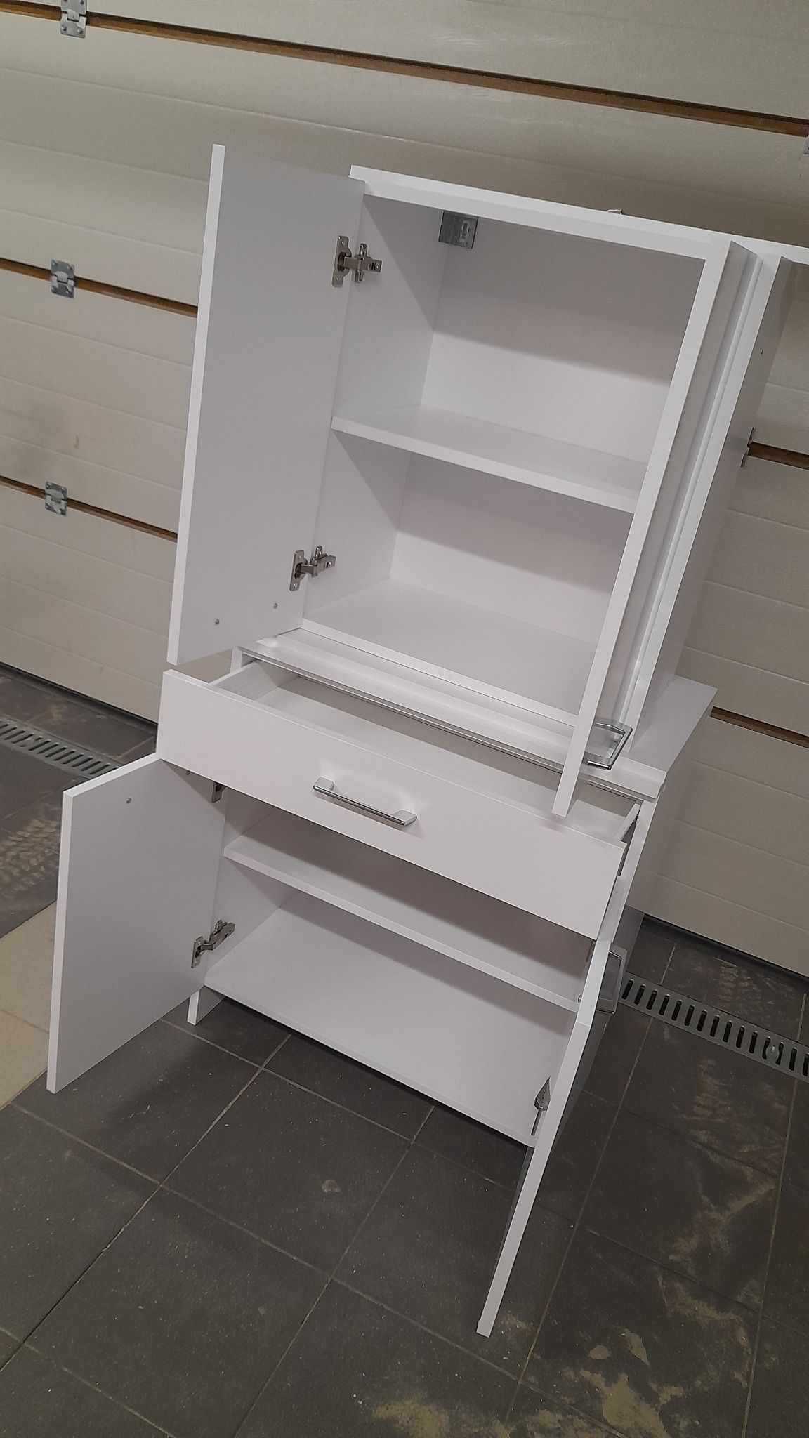 Biała szafka z otwartymi drzwiczkami i szufladą, widok z boku na tle garażowej bramy. Prosta forma, minimalistyczny design, metalowe uchwyty.