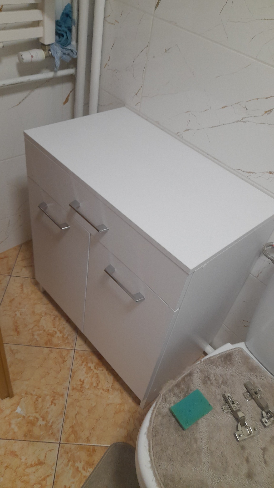 Biała szafka łazienkowa z dwoma drzwiczkami i srebrnymi uchwytami, stojąca na brązowych płytkach, widok z góry, elementy montażowe na sedesie.