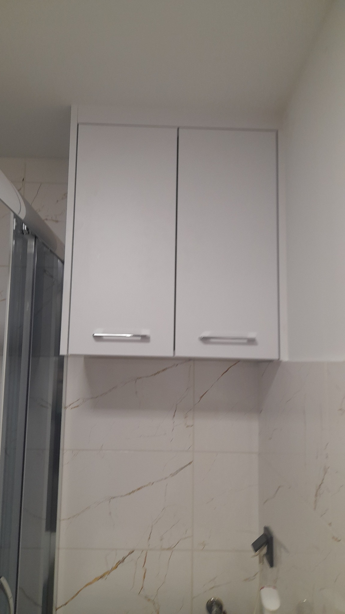 Biała szafka łazienkowa z dwoma drzwiczkami i srebrnymi uchwytami, zamontowana nad umywalką z marmurowymi kafelkami. Widoczny fragment kabiny prysznicowej.