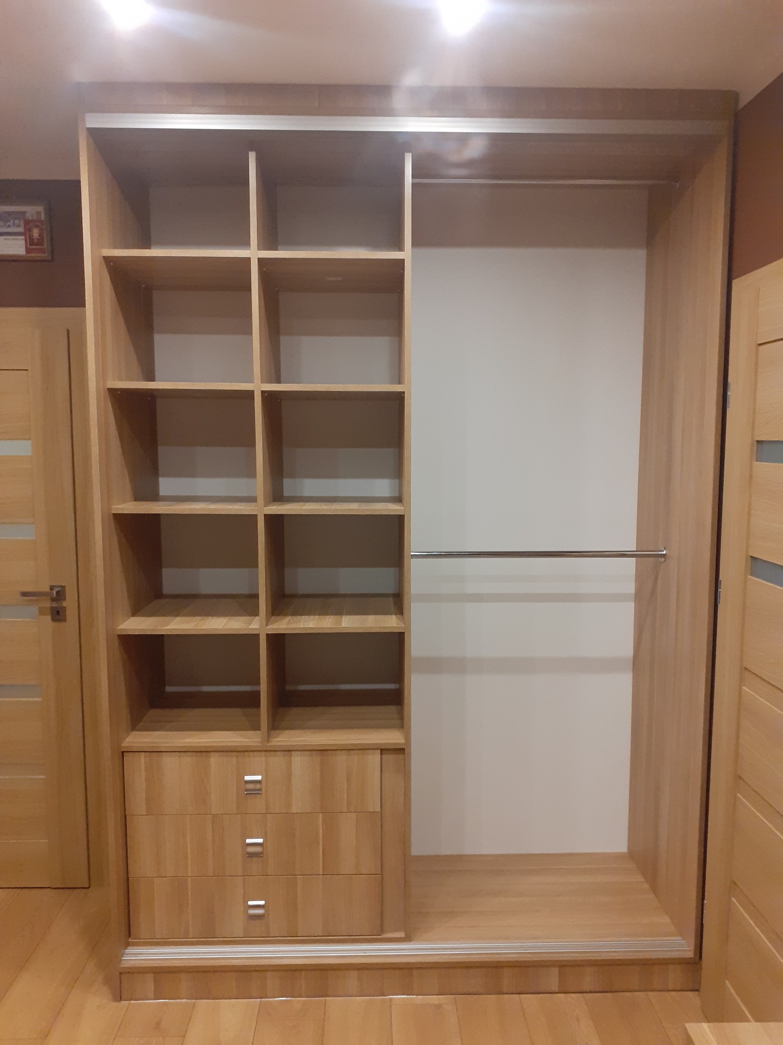 Jasna, drewniana szafa wnękowa z półkami i szufladami, widoczna na tle podłogi z paneli, z drzwiami po prawej stronie. Wnętrze szafy jest puste.